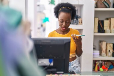 African american woman shop assistant using computer talking on smartphone at clothing store