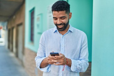 Genç Arap adam sokakta akıllı telefon kullanarak kendinden emin gülümsüyor.