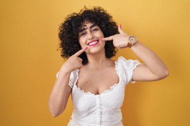 Sarı arka planda kıvırcık saçlı, neşeli gülümseyen ve parmaklarıyla ve ağzıyla işaret eden genç esmer kadın. diş sağlığı kavramı. 