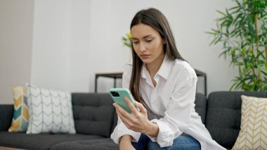 Genç, güzel, İspanyol bir kadın evde ciddi bir ifadeyle akıllı telefon kullanıyor.