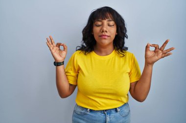 Mavi arka planda duran İspanyol kadın rahatlamış ve gözleri kapalı gülümsüyor parmaklarıyla meditasyon hareketi yapıyor. yoga konsepti. 