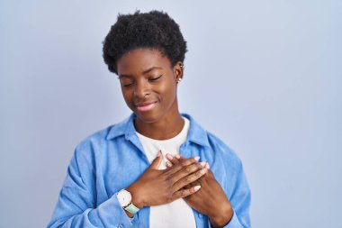 African american woman standing over blue background smiling with hands on chest with closed eyes and grateful gesture on face. health concept. 