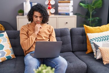 Evdeki kanepede dizüstü bilgisayar kullanan genç latin adam.