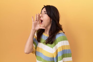 Orta yaşlı İspanyol bir kadın sarı arka planda durup el pençe divan durup el ele bağırıyor. iletişim konsepti. 