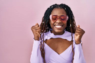 Pembe arka planda örülü saçları olan Afrikalı kadın kolları havada ve gözleri kapalı zaferi kutlarken heyecanla bekliyor. Kazanan konsept. 