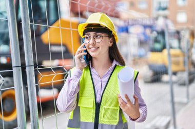 Young hispanic woman architect talking on the smartphone holding blueprints at street