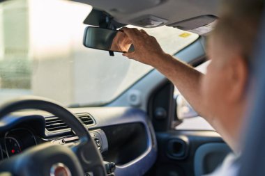 Orta yaşlı, gri saçlı bir adam dikiz aynasına dokunuyor. Sokakta arabada oturuyor.