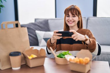 Genç bir kadın akıllı telefonuyla fotoğraf çektirip evdeki yiyecekleri alıyor.