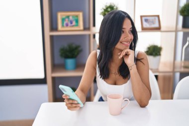 Young hispanic woman drinking coffee and using smartphone at home