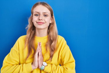 Mavi arka planda dikilen beyaz bir kadın ellerini birleştirip af diliyor kendinden emin bir şekilde gülümsüyor.. 