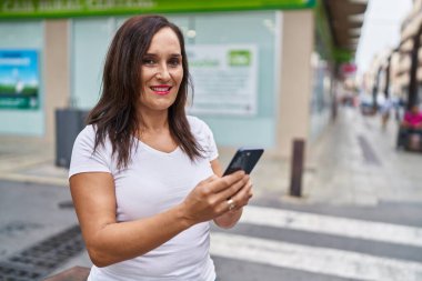 Genç, güzel, İspanyol bir kadın sokakta akıllı telefon kullanarak gülümsüyor.