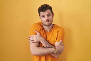 Young hispanic man standing over yellow background shaking and freezing for winter cold with sad and shock expression on face 