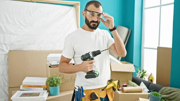 若いヒスパニックの男性修理マンは 新しい家でセキュリティメガネを着用ドリルを保持しています — ストック写真