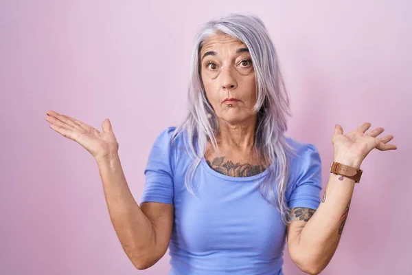 ピンクを背景にしたタトゥーのある中年女性は 腕や手を上げて混乱した表情を見せていました 疑わしい概念 — ストック写真