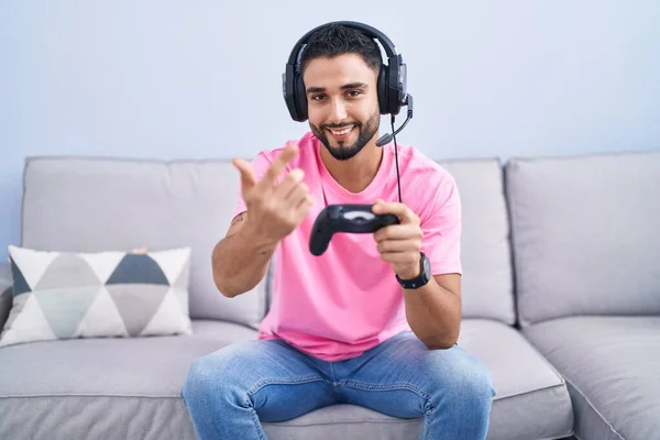 西班牙裔年轻人坐在沙发上玩电子游戏 手握控制器 招手欢迎 微笑着来到这里 — 图库照片
