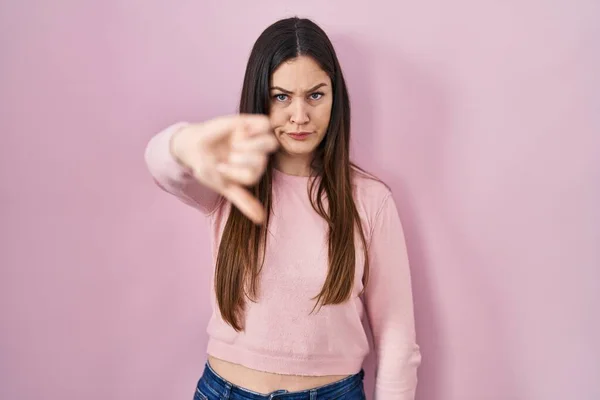 ピンクの背景に立っている若いブルネットの女性は 親指を下にジェスチャーで拒絶と否定を示す不満と怒りを見ています 悪い表現 — ストック写真