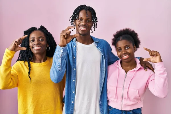 一组三个年轻的黑人站在粉红的背景上 微笑着 自信地做手势 手拿着小尺寸的标志 手拿着手指 手拿着相机 计量概念 — 图库照片