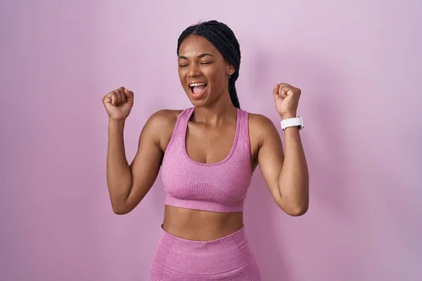 ピンクの背景にスポーツウェアを身に着けているアフリカ系アメリカ人女性は 腕を上げて目を閉じて成功に驚いて驚いています 勝者のコンセプト — ストック写真
