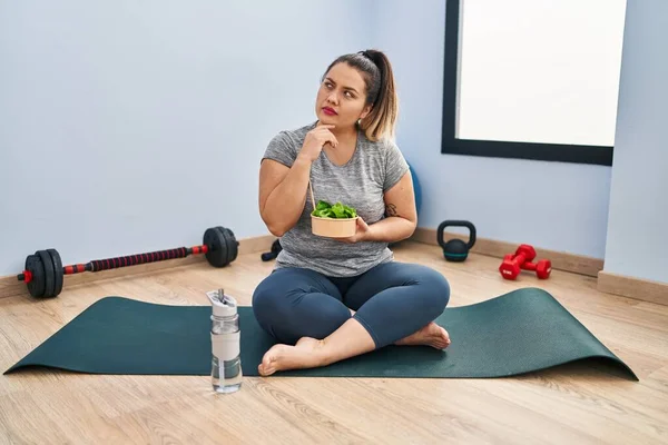 年轻的惊慌失措的女人坐在瑜伽垫上吃着健康的沙拉严肃的脸 手托着下巴思考着问题 思考着困惑的想法 — 图库照片