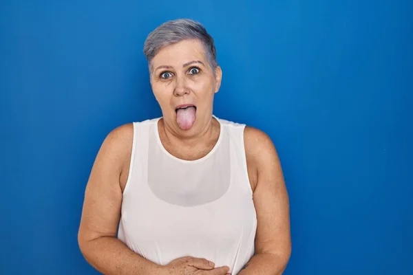 中年白人女人站在蓝色的背景上 带着滑稽的表情伸出舌头 高兴极了 情感概念 — 图库照片