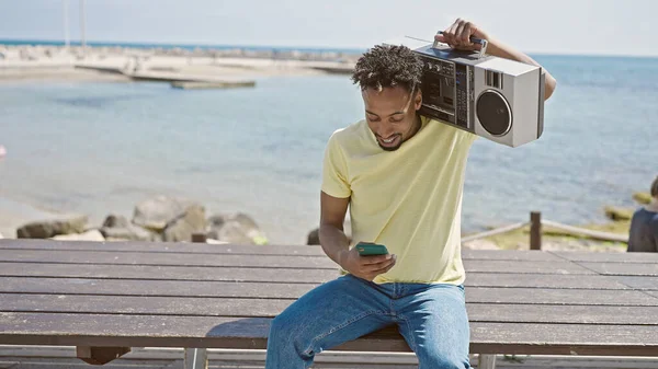 アフリカ系アメリカ人男性が海辺でスマートフォンを使ってブームボックスを持っている — ストック写真