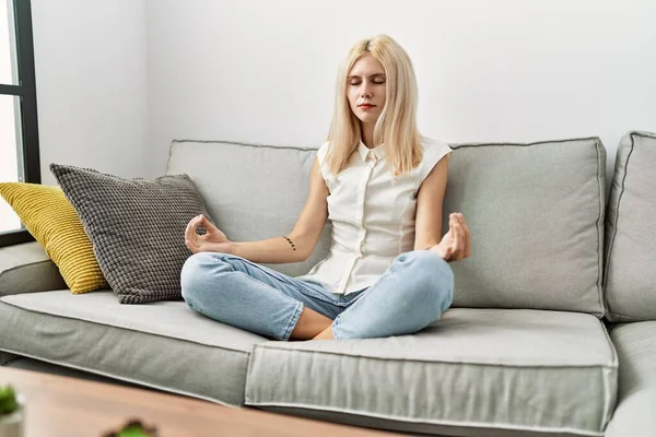 年轻的金发女子坐在家里的沙发上做瑜伽运动 — 图库照片