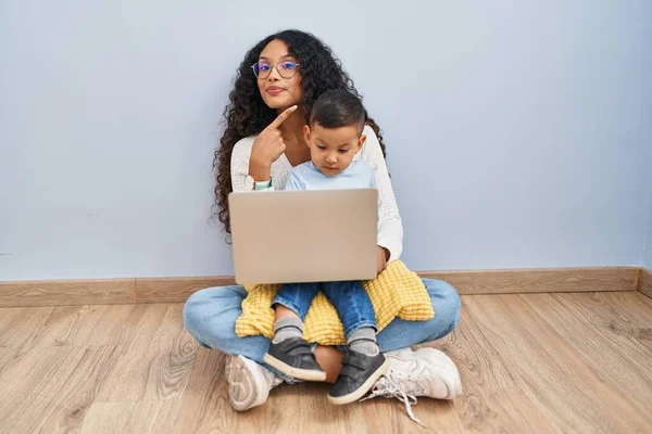 深刻で穏やかな顔を示す側面への手の指で指して床に座ってコンピュータのラップトップを使用して若いヒスパニック系の母親と子供 — ストック写真