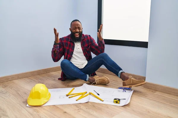 アフリカ系アメリカ人の男性が床に新しい家で腕を上げ 興奮して叫んで目を閉じて成功のために狂気と狂気を祝う設計図を見て座っている 勝者のコンセプト — ストック写真