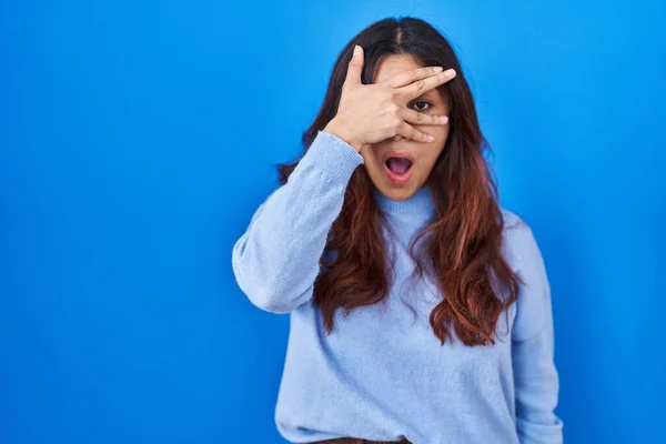 stock image Hispanic young woman standing over blue background peeking in shock covering face and eyes with hand, looking through fingers with embarrassed expression. 