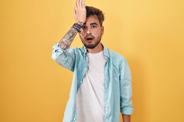 Young hispanic man with tattoos standing over yellow background surprised with hand on head for mistake, remember error. forgot, bad memory concept. 