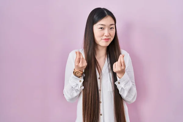 ピンクの背景に立っている中国の若い女性は手でお金のジェスチャーをし 給与支払いを求め 億万長者のビジネス — ストック写真