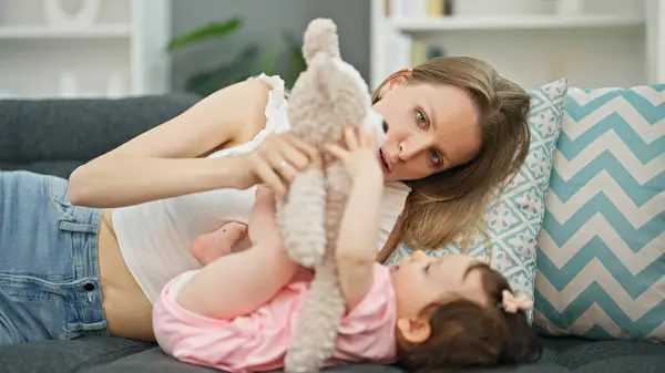 Mother and daughter lying on sofa playing with teddy bear at home
