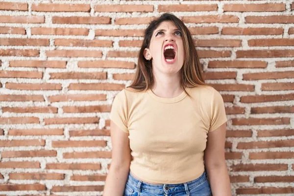 若いブルネットの女性がレンガの壁の上に立って怒りと怒りで叫んで イライラし 激怒叫んで怒っている 怒りと攻撃的な考え方 — ストック写真