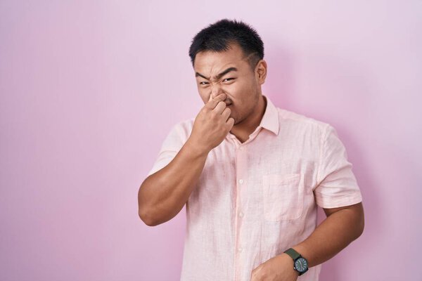 Chinese young man standing over pink background smelling something stinky and disgusting, intolerable smell, holding breath with fingers on nose. bad smell 