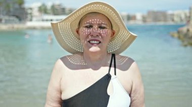 Orta yaşlı, gri saçlı, mayo giyen, yaz şapkalı, kumsalda öpüşen bir kadın turist.