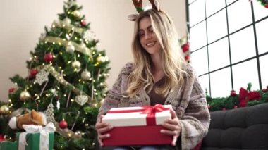 Sarışın bir kadın hediye paketini açıyor evdeki Noel ağacının yanındaki koltukta oturuyor.