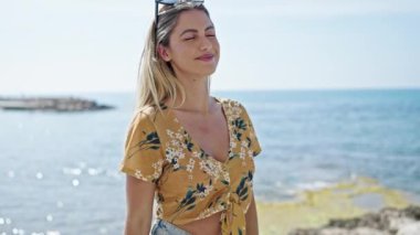 Genç sarışın kadın gülümsüyor. Kollarını çapraz yapıp sahilde duruyor.