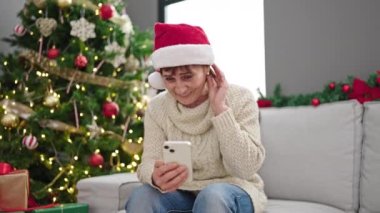 Evde Noel ağacının yanında oturan akıllı telefon kullanan olgun İspanyol kadın.