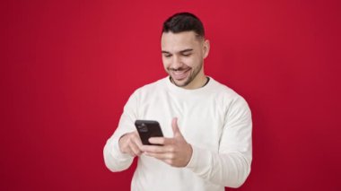 Genç İspanyol adam, izole edilmiş kırmızı arka planda gülümseyen akıllı telefon kullanıyor.