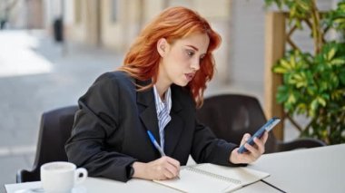 Genç kızıl saçlı iş kadını akıllı telefon kullanıyor. Kahve dükkanının terasında not alıyor.