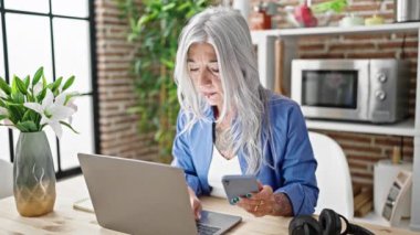 Orta yaşlı, gri saçlı, dizüstü bilgisayar ve akıllı telefon kullanan bir kadın yemek odasındaki masada oturuyor.