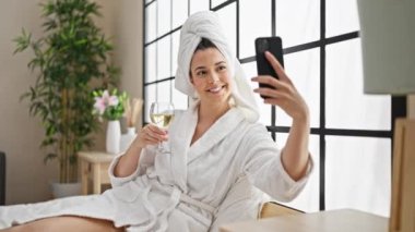 Bornoz giymiş, şampanya içen genç ve güzel bir İspanyol kadın yatak odasında akıllı telefonlardan selfie çekiyor.