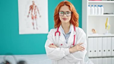Genç kızıl saçlı kadın doktor gülümsüyor. Klinikte kollarını kavuşturup duruyor.