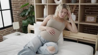 Genç hamile kadın yatak odasında gergin bir ifadeyle karnına dokunuyor.