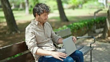 Genç İspanyol adam parkta bankta dizüstü bilgisayar kullanıyor.