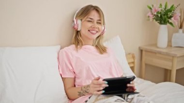 Young blonde woman watching video sitting on bed at bedroom