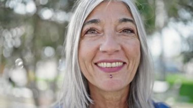 Orta yaşlı, kır saçlı, gülümseyen, kendine güvenen bir kadın parkta duruyor.