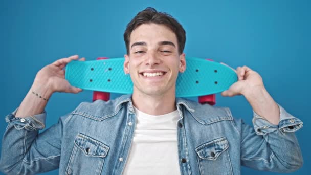 年轻的他的惊慌失措的男人带着自信的笑容 把滑板举过孤独的蓝色背景 — 图库视频影像