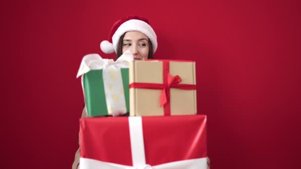 クリスマスの帽子をかぶった美しいヒスパニックの女性は 孤立した赤い背景に贈り物を保持します — ストック動画