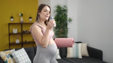 Evde su içen yoga minderi tutan hamile bir kadın.